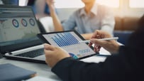 Two investors discussing ROI with graphs displayed on a tablet and a laptop